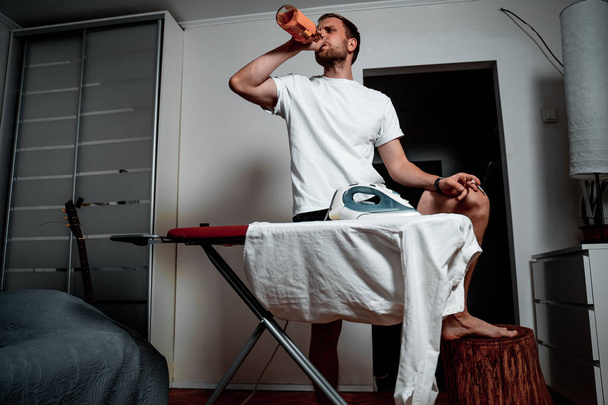 A man in a white T-shirt with a cigarette and a drink ironing his shirt - Photo, Image