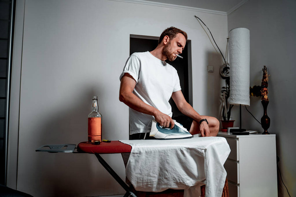 A man in a white T-shirt with a cigarette and a drink ironing his shirt - Fotoğraf, Görsel