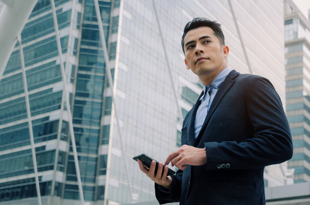 young handsome asian business man wearing modern black suit reading information about finance news with mobile smart phone in building city background, network technology, internet, successful concept - Foto, afbeelding