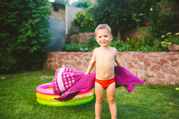 Thème vacances d'été. Un petit garçon caucasien de 3 ans jouant dans la cour d'une maison sur l'herbe près d'une piscine gonflable ronde de couleur enveloppée, essuyée pour être une serviette sèche dans des troncs de natation rouges
. - Photo, image