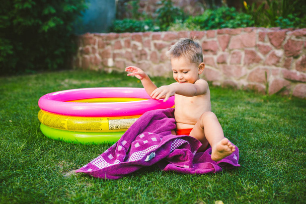 Summer vacation theme. A small 3 year old Caucasian boy playing in the backyard of a house on the grass near a round inflatable colored pool wrapped up, wiped to be a dry towel in red swimming trunks. - Valokuva, kuva