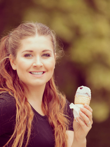 Happy funny young woman with long brown hair eating ice cream having fun enjoying her dessert during beautiful summer weather against green park - Foto, Imagem