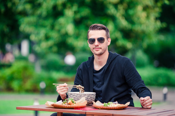 Портрет молодої людини, що їсть азіатську вуличну їжу з паличками та супом для хутра у відкритому кафе
 - Фото, зображення