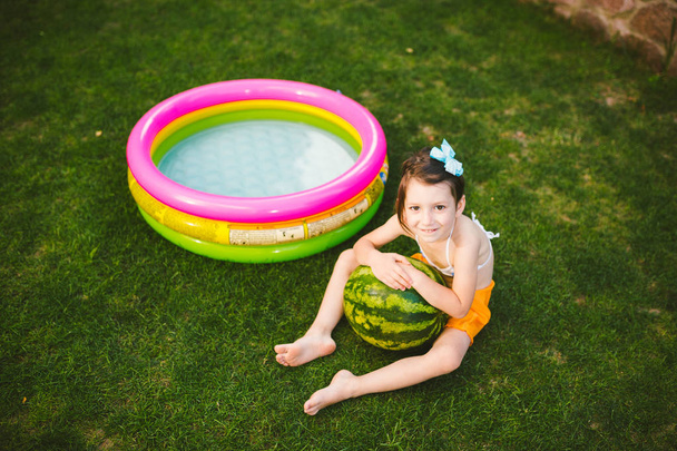 The topic proper healthy eating children. Little baby girl 4 years old caucasian sitting on green grass hugging big green round berries watermelon near childrens inflatable pool in yard of house - Photo, Image