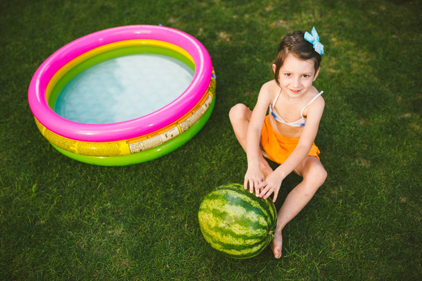 das Thema gesunde Ernährung von Kindern. kleines Mädchen 4 Jahre alt Kaukasier sitzt auf grünem Gras umarmt große grüne runde Beeren Wassermelone in der Nähe von Kindern aufblasbaren Pool im Hof des Hauses - Foto, Bild