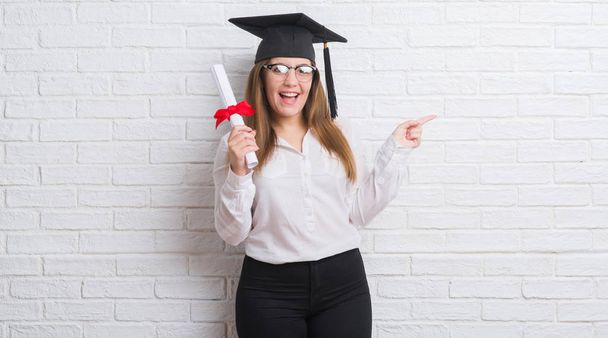 Jeune femme adulte sur mur de briques blanches portant chapeau diplômé tenant degré très heureux pointant avec la main et le doigt sur le côté
 - Photo, image