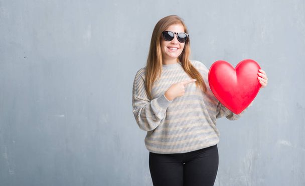 Молодая взрослая женщина над серой гранж-стеной держа красное сердце в любви очень счастливо указывая рукой и пальцем
 - Фото, изображение