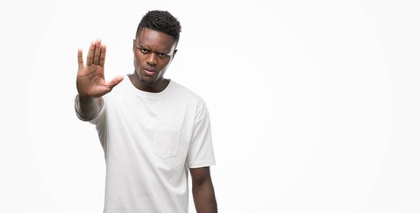 Молодой африканский американец в белой футболке делает остановку петь с ладонью руки. Предупреждающее выражение с негативным и серьезным жестом на лице
. - Фото, изображение