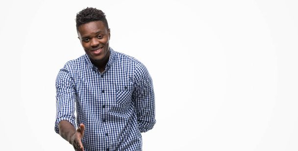Νεαρός αφρικανική αμερικανική άνθρωπος που φοράει μπλε πουκάμισο χαμογελώντας προσφέροντας φιλική χειραψία ως χαιρετισμό και καλωσόρισμα. Επιτυχημένη επιχείρηση. - Φωτογραφία, εικόνα