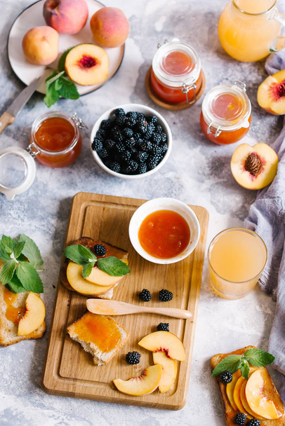 Toasts of bread with apricot jam and fresh fruits with leaves on white wooden table. Tasty breakfast. - Foto, Bild