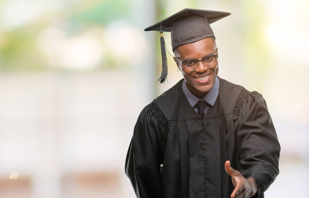 Joven hombre afroamericano graduado sobre fondo aislado sonriendo amistoso ofreciendo apretón de manos como saludo y bienvenida. Negocios exitosos
. - Foto, imagen
