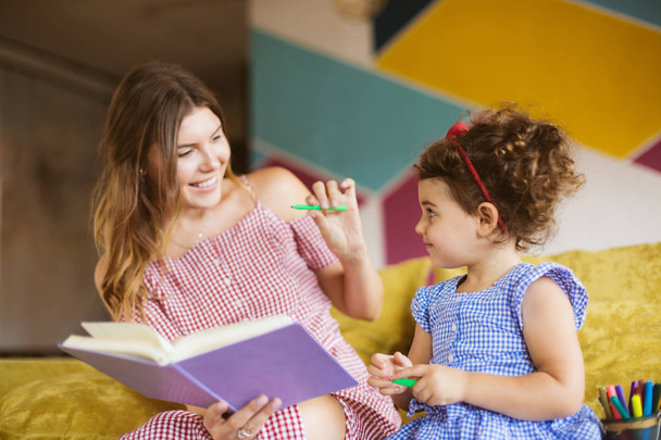 schöne lächelnde Mutter mit ihrer kleinen süßen Tochter, die fröhlich zusammen auf dem heimischen Sofa Kinderbücher liest - Foto, Bild