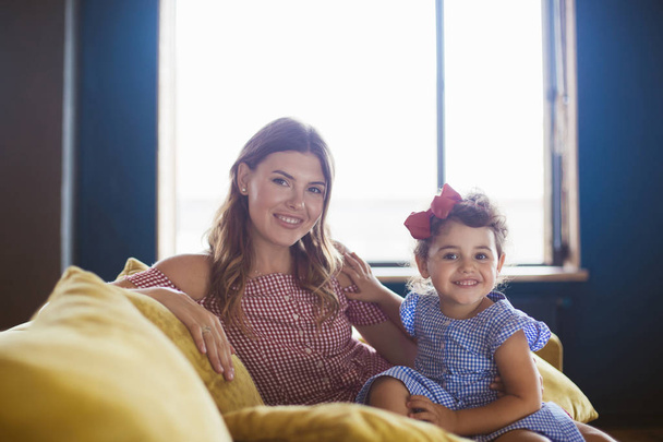 Beautiful smiling mother with her little cute daughter in blue dress happily looking in camera spending time together on sofa at home with big window on background - Photo, Image