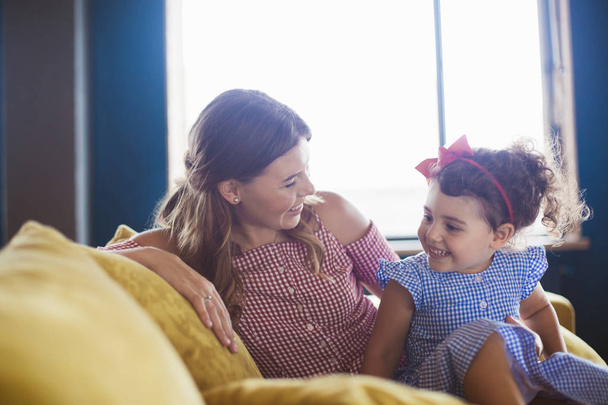 Hermosa madre sonriente con su pequeña hija bonita en vestido azul con alegría pasar tiempo juntos en el sofá en casa
 - Foto, imagen