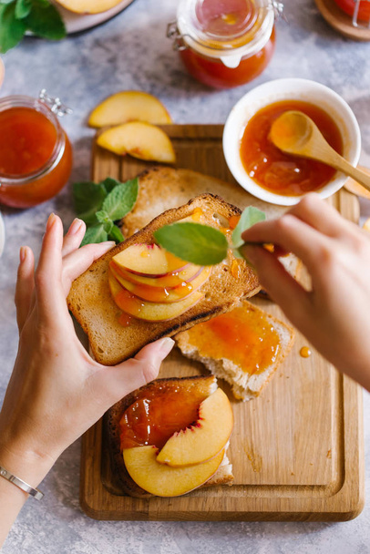 Toasts of bread with apricot jam and fresh fruits with leaves on white wooden table. Tasty breakfast. - Foto, afbeelding