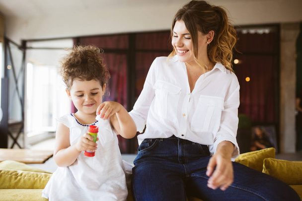 Joven madre sonriente en camisa blanca con la pequeña hija bonita en vestido alegremente soplando burbujas pasar tiempo juntos en casa
  - Foto, imagen