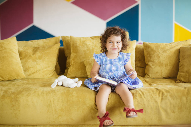 Cute little smiling girl with dark curly hair in blue dress holding book in hand dreamily looking aside on sofa at home  - Photo, Image