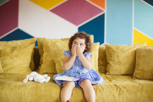 Cute little girl with dark curly hair in blue dress with child book on knees dreamily looking aside covering mouth with hands on sofa at home  - Foto, afbeelding
