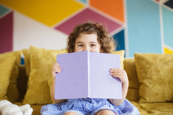 Bella bambina con i capelli ricci scuri in abito blu felicemente alla ricerca in bocca fotocamera che copre con libro sul divano a casa
 - Foto, immagini