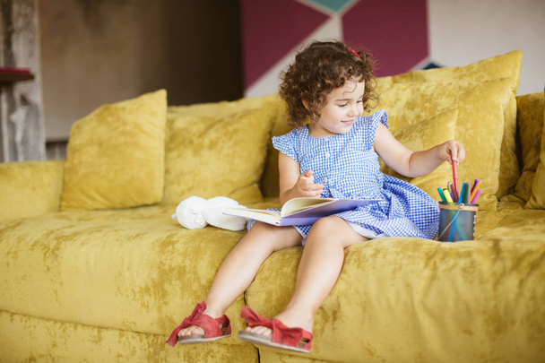 Ładna dziewczynka uśmiechający się ciemne, kręcone włosy w niebieskiej sukience z książki na kolana wybór kolorowych flamastrów na kanapie w domu  - Zdjęcie, obraz