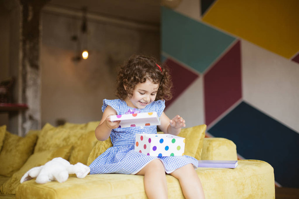Joyful baby girl with dark curly hair in blue dress happily opening gift box on sofa at home  - Photo, Image