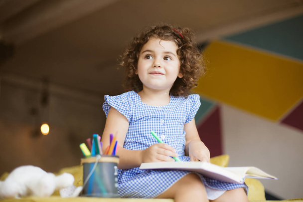 Belle petite fille aux cheveux bouclés foncés en robe bleue regardant de côté le dessin dans un livre à colorier avec des stylos à pointe de feutre sur le canapé à la maison
  - Photo, image