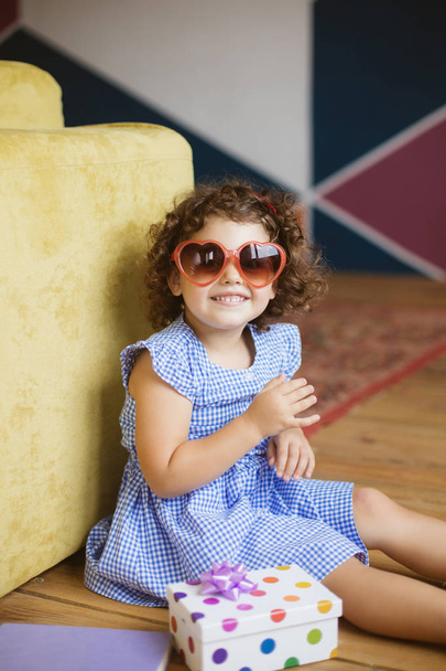 Linda niña sonriente con pelo rizado oscuro en vestido azul y gafas de sol mirando felizmente en la cámara con caja de regalo en el suelo en casa
  - Foto, imagen