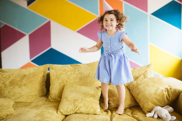hübsches fröhliches kleines Mädchen mit dunklem lockigem Haar in blauem Kleid und Geburtstagskappe, das zu Hause fröhlich auf dem Sofa hüpft  - Foto, Bild