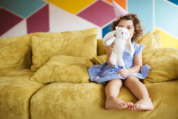 Beautiful baby girl with dark curly hair in blue dress and birthday cap happily showing rabbit toy on camera on sofa at home  - Foto, imagen