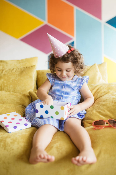 Bastante niña sonriente con pelo rizado oscuro en vestido azul y gorra de cumpleaños felizmente apertura caja de regalo en el sofá en casa
 - Foto, Imagen
