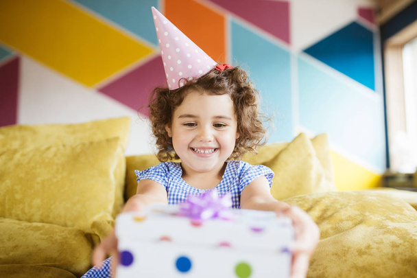 Iloinen pieni tyttö, jolla on tummat kiharat hiukset syntymäpäiväkorkissa onnellisesti pitämällä lahjapakkausta käsissä sohvalla kotona
 - Valokuva, kuva