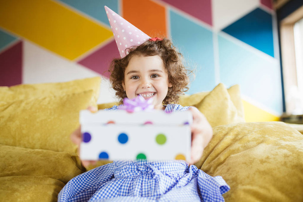 Hermosa niña sonriente con cabello rizado oscuro en gorra de cumpleaños mirando felizmente en la cámara sosteniendo la caja de regalo en las manos en el sofá en casa
 - Foto, imagen