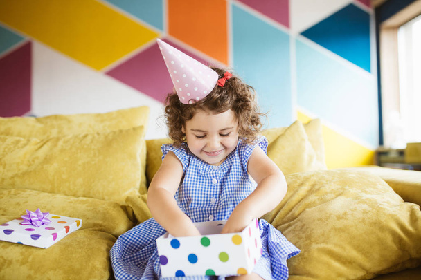 Ładny uśmiechający się mała dziewczynka, ciemne kręcone włosy w urodziny cap szczęśliwie otwierając pudełko na kanapie w domu - Zdjęcie, obraz