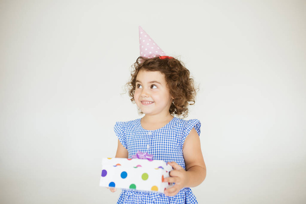 hübsches lächelndes Baby-Mädchen mit dunklem lockigem Haar in blauem Kleid und Geburtstagskappe, das fröhlich zur Seite schaut und Geschenkschachtel über weißem Hintergrund hält - Foto, Bild