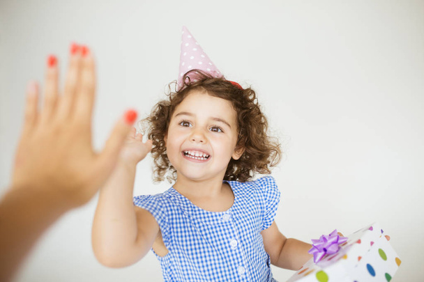 Smilig meisje met donkere krullend haar in blauwe jurk en verjaardag cap gelukkig hoog geven vijf met gift box in hand op witte achtergrond - Foto, afbeelding