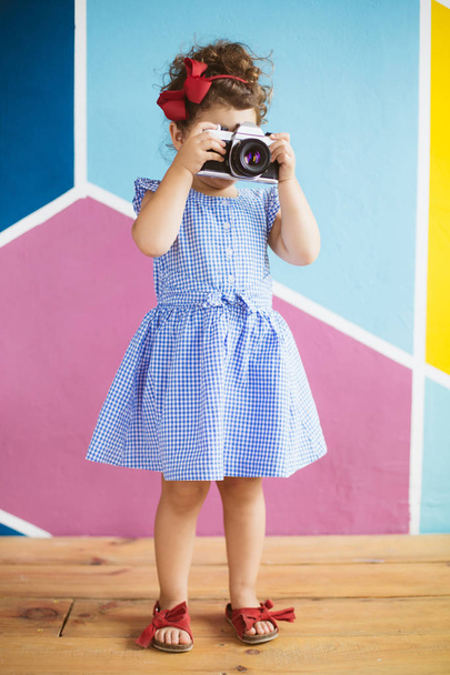 Cute little girl with dark curly hair in blue dress playfully covering face with retro camera over colorful background - Photo, image
