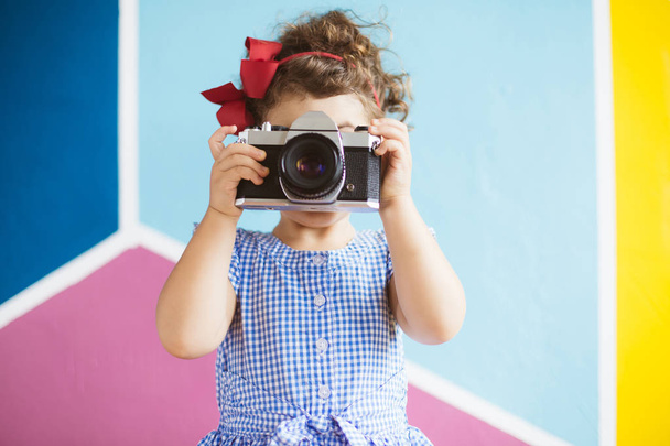 Pretty little girl with dark curly hair in dress playfully covering face with retro camera over colorful background - Foto, Imagen