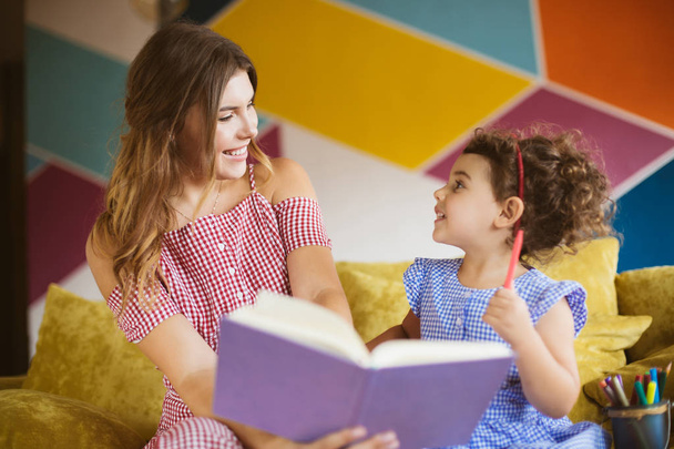 Hermosa madre sonriente con su pequeña hija linda felizmente mirándose el uno al otro leyendo libro infantil juntos en el sofá en casa
 - Foto, imagen