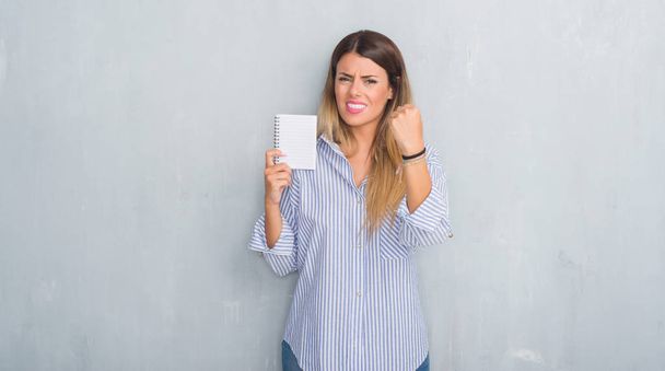 Young adult woman over grey grunge wall holding blank notebook annoyed and frustrated shouting with anger, crazy and yelling with raised hand, anger concept - Foto, immagini