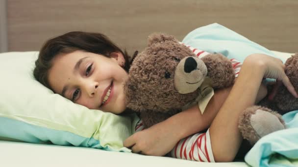 Too tired today. Cute happy young girl lying in her bad hugging her teddy bear toy smiling to the camera falling asleep children kids childhood happiness sleepy sleep relaxation resting rest concept - Imágenes, Vídeo