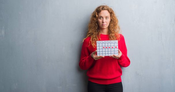 Молодая рыжая женщина через серый гранж стены холдинга календарь период с уверенным выражением на умное лицо думает серьезно
 - Фото, изображение