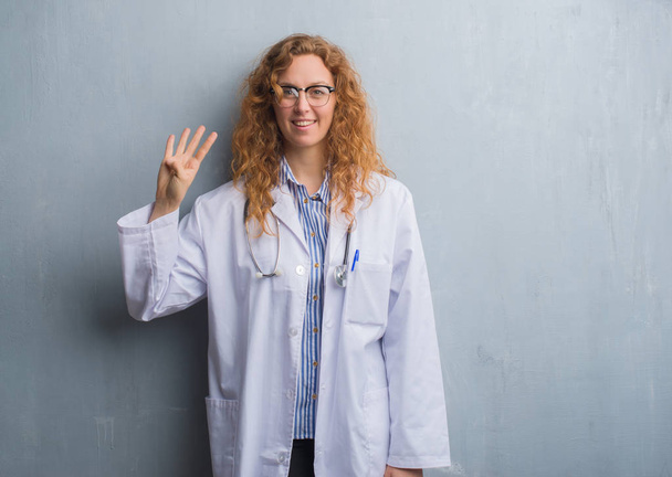junge rothaarige Ärztin über grauer Grunge-Wand in einem Mantel, der Finger Nummer vier zeigt und nach oben zeigt, während sie selbstbewusst und glücklich lächelt. - Foto, Bild
