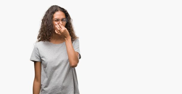 Beautiful young hispanic woman wearing glasses smelling something stinky and disgusting, intolerable smell, holding breath with fingers on nose. Bad smells concept. - Foto, afbeelding