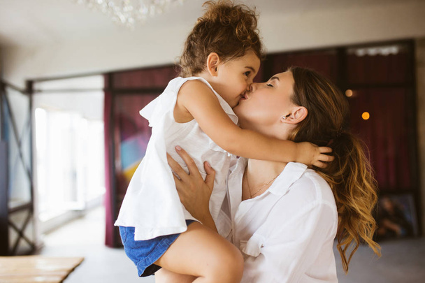 Beautiful young mother in white shirt holding little cute daughter in hands and kissing spending time together at home - Photo, Image