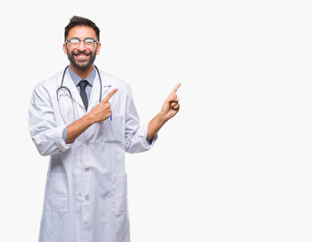 Médico hispano adulto sobre fondo aislado sonriendo y mirando a la cámara apuntando con dos manos y dedos hacia un lado
. - Foto, imagen