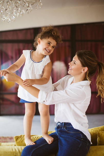 Nice young mother in white shirt and jeans happily holding little smiling daughter on knees spending time together at home - Photo, Image