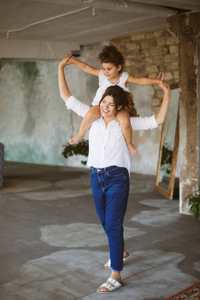Young beautiful mother in white shirt and jeans holding little cute daughter on shoulders dreamily spending time together at home - Photo, Image