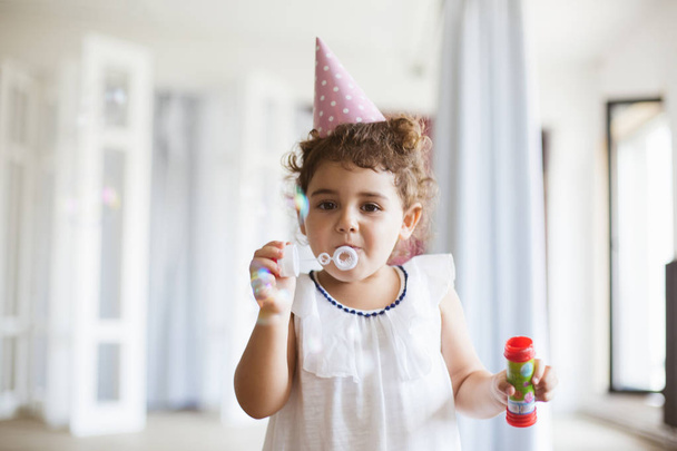 Beautiful little girl with dark curly hair in dress and birthday cap dreamily looking in camera blowing bubbles at home - Photo, Image