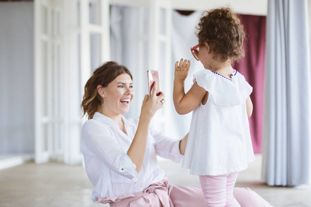Веселая мама радостно фотографирует на мобильный телефон симпатичную маленькую дочь в очках для взрослых, проводящую время вместе дома
 - Фото, изображение
