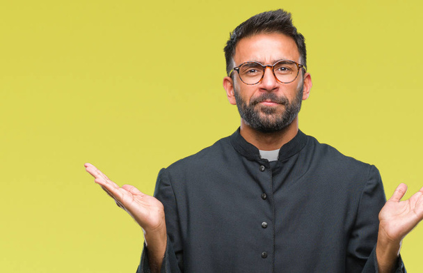 Erwachsener hispanischer katholischer Priester Mann mit isoliertem Hintergrund, ahnungsloser und verwirrter Miene mit erhobenen Armen und Händen. Zweifelhaftes Konzept. - Foto, Bild